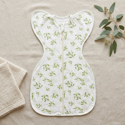 Newborn asleep on back in the CradleUp arms-up swaddle, green leaf print, in a sunlit nursery