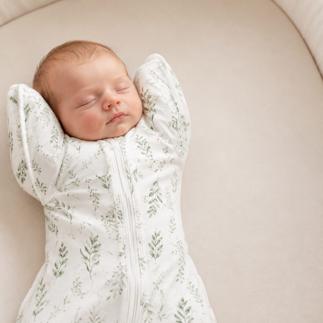 Newborn sleeping peacefully with arms up in the CradleUp swaddle, sage-green leaf print